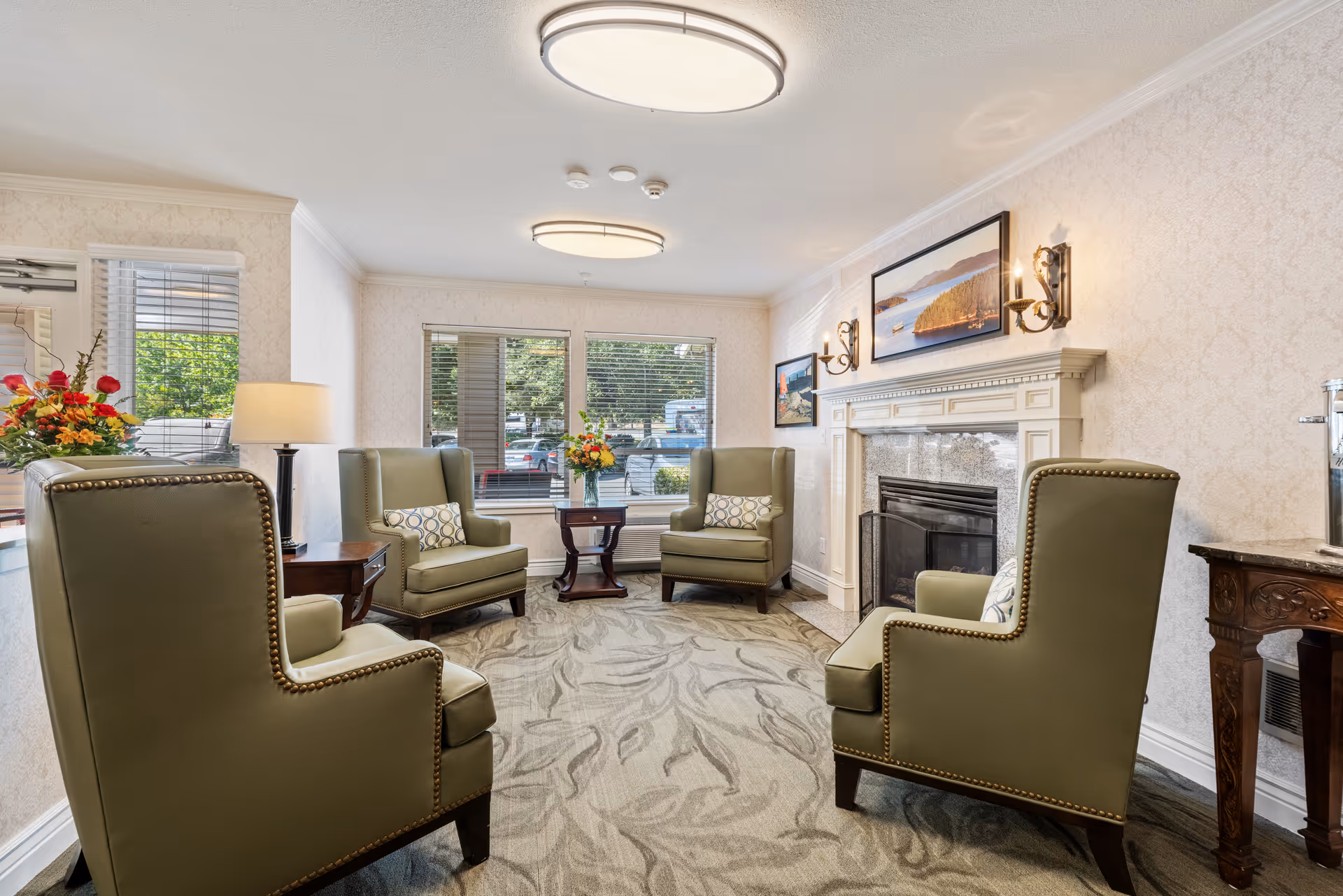 Bright communal sitting area with four green wingback chairs arranged around a fireplace and windowed wall.