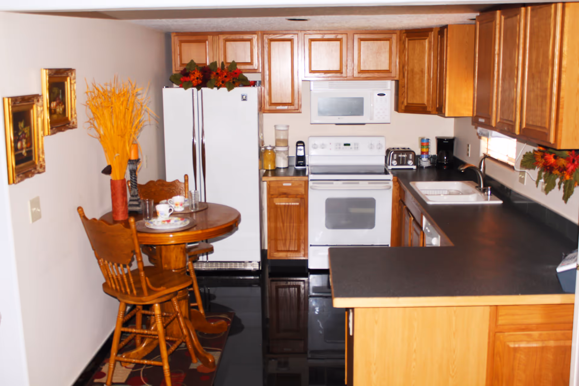 A kitchen area with wooden cabinets, a white refrigerator, a white stove with an oven, a white microwave above the stove, and a black countertop with a sink. There is a small round wooden table with two wooden chairs, decorated with a vase of yellow dried flowers and a tea set. The walls have framed pictures and a decorative garland near the window.