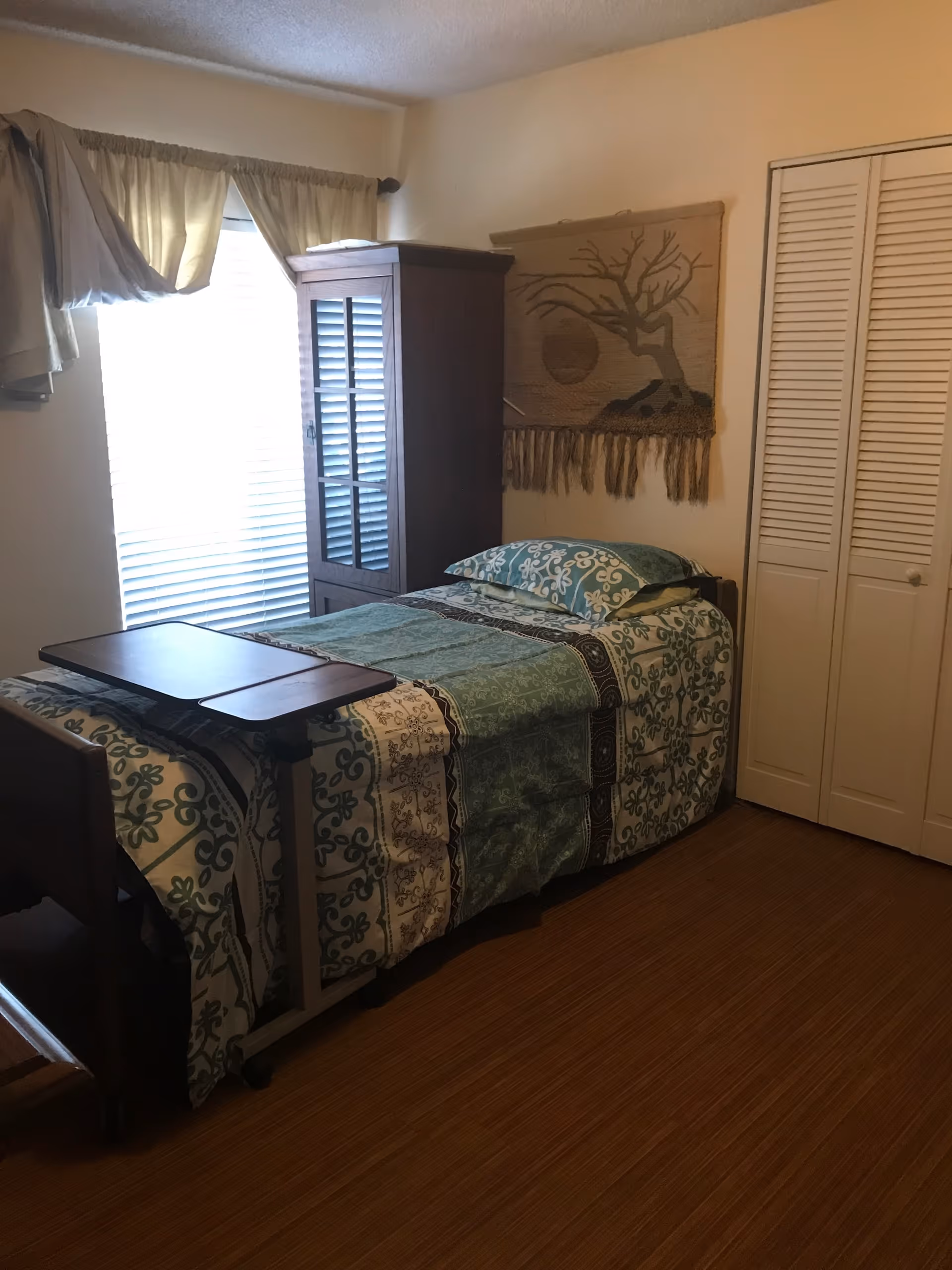 A small bedroom with a single bed covered in a patterned green and white bedspread. There is a wooden overbed table positioned over the bed. Next to the bed is a tall wooden cabinet with glass doors. A window with blinds and beige curtains is on the left wall. On the right wall, there is a woven wall hanging depicting a tree and sun, and a closed white closet door. The floor is wooden.
