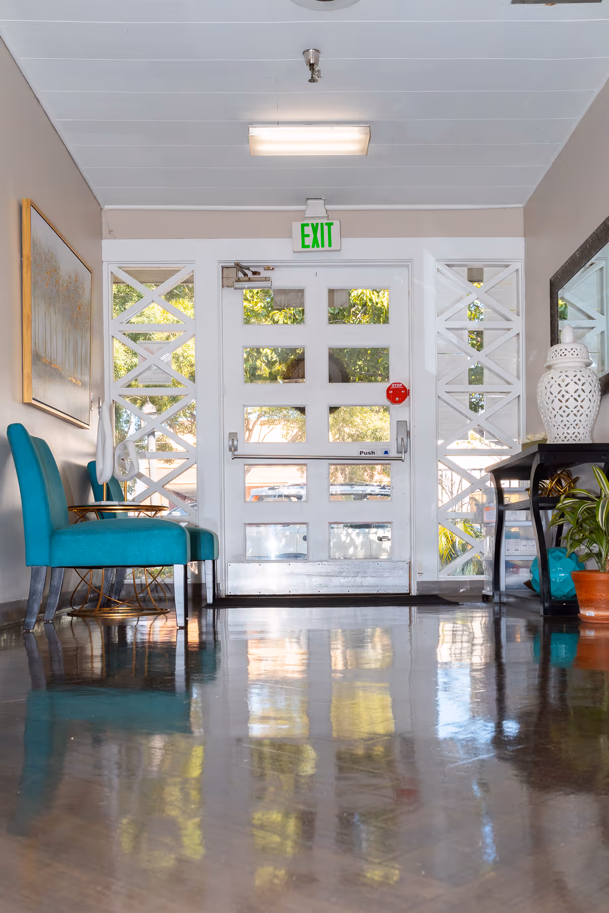 Interior view of a hallway or entrance area with a white door featuring multiple glass panels and an illuminated green exit sign above it. On the left side, there are two teal upholstered chairs and a small round table. On the right side, there is a black console table with decorative items including a white ceramic vase and a potted plant. The floor is shiny and reflective.