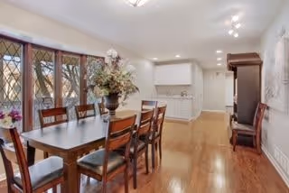 Bright dining room with a long wooden table and chairs, a floral centerpiece, hardwood floors, and a kitchen area visible in the background.