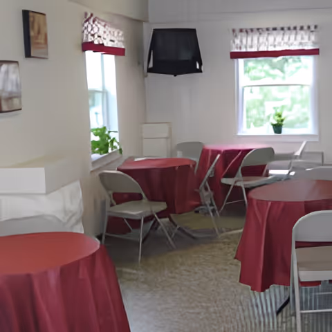 Small dining/activity room featuring round tables with red tablecloths, folding chairs, windows with valances, and a wall-mounted TV.