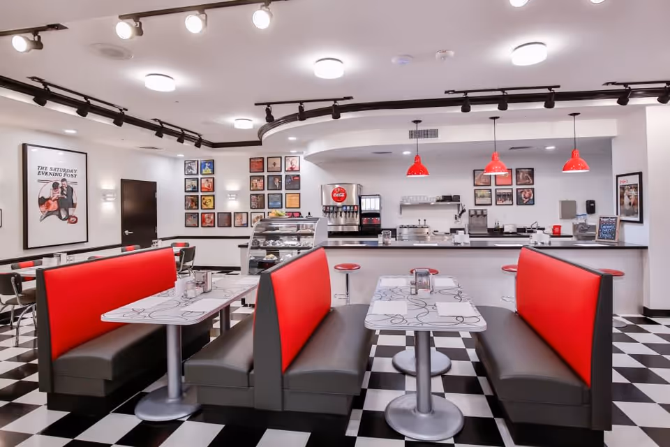 A retro-style dining area with black and white checkered floor, red and gray booth seating, and tables with white tops featuring black swirl designs. The back wall has a counter with red pendant lights hanging above, a soda fountain, and a display case with pastries. The walls are decorated with framed pictures and posters, including a large 'The Saturday Evening Post' cover on the left.