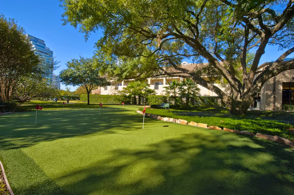 Outdoor putting green with several small red flags, surrounded by trees and bushes, with a building and a tall city building visible in the background under a clear blue sky.