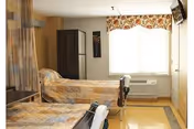 A small, clean bedroom with two single beds covered in beige and brown patterned bedspreads. There is a window with a floral valance letting in natural light, a dark wooden wardrobe, and a wall-mounted television. The room has light-colored walls and a yellowish floor.