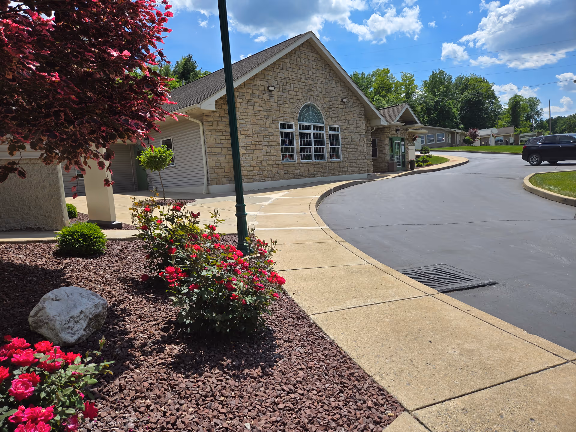 Front entrance of a stone-faced senior care building with a curved driveway, landscaped flower beds, and a lamp post.