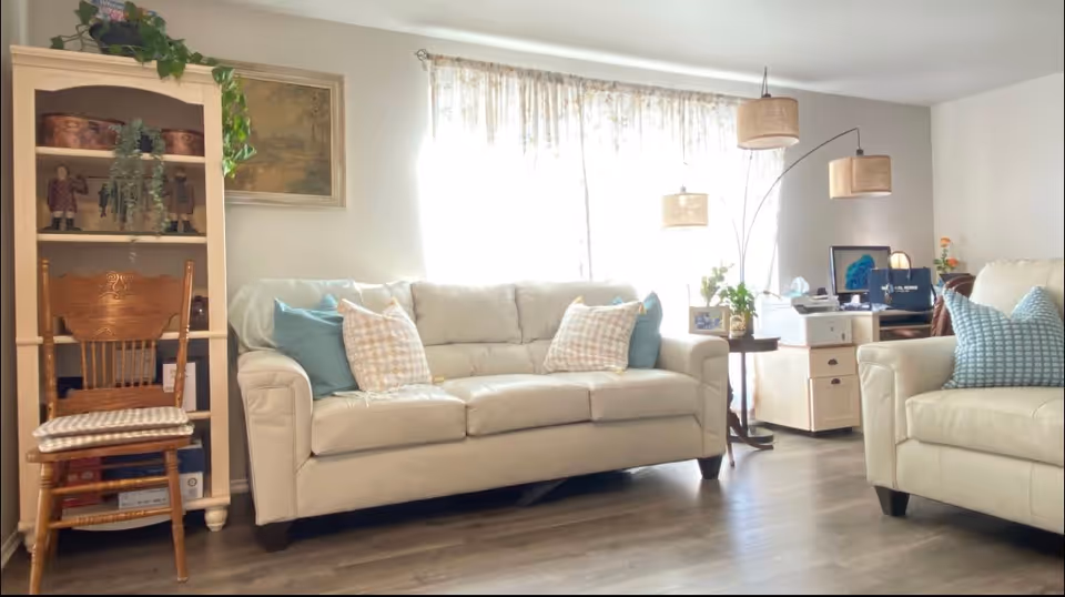 A bright living room with two cream-colored leather sofas adorned with blue and patterned throw pillows. There is a wooden chair with a cushioned seat next to a white shelving unit holding decorative items and plants. A floor lamp with three beige lampshades stands near a small side table with framed photos and a plant. A desk with a computer and office supplies is visible in the background. The room has light-colored walls and wood flooring, with sheer curtains allowing natural light to fill the space.