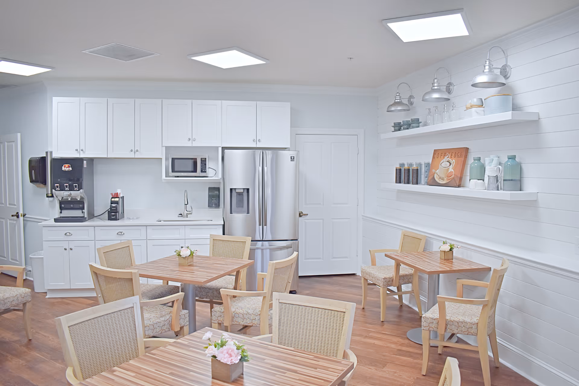 A bright and clean kitchen and dining area with white cabinets, a stainless steel refrigerator, a microwave, and a coffee machine. There are several wooden tables with cushioned chairs arranged around the room, each table decorated with a small flower arrangement. White shelves on the wall hold jars, cups, and a coffee-themed sign. The floor is wooden, and the room is well-lit with ceiling lights.