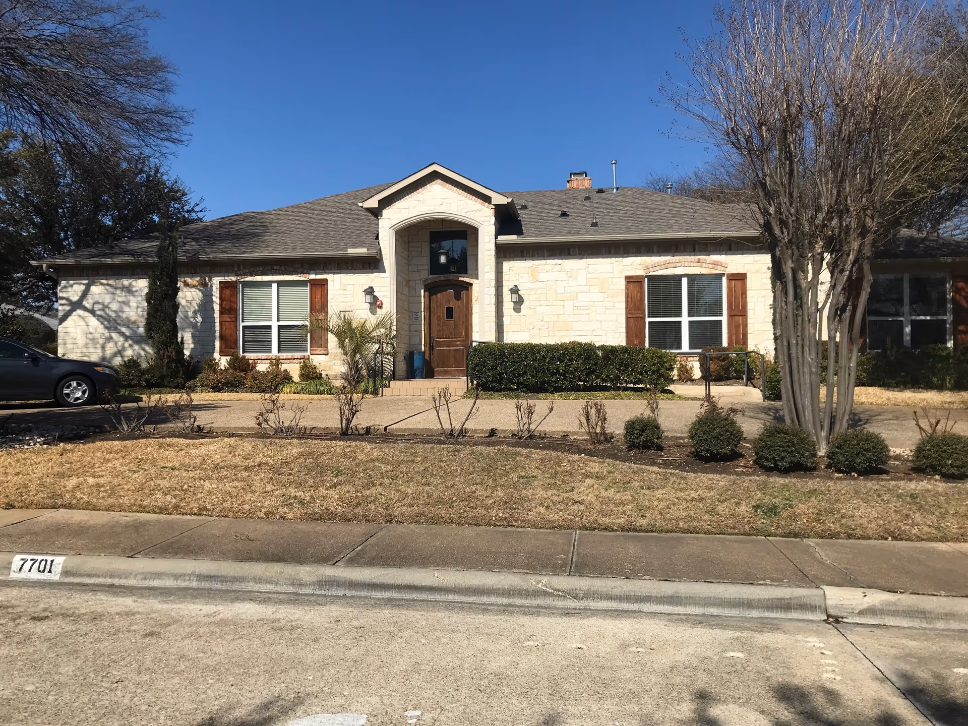 Front exterior view of a single-story stone building with a gabled roof, wooden front door, and windows with wooden shutters. There is a small landscaped area with bushes and trees in front of the building, a sidewalk, and a parked car to the left. The sky is clear and blue.