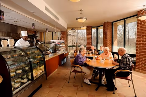 A group of elderly people sitting around a round table in a well-lit dining area with large windows. A chef stands behind a glass display case filled with pastries and baked goods. The room has tiled floors, brick walls, and pendant lighting.