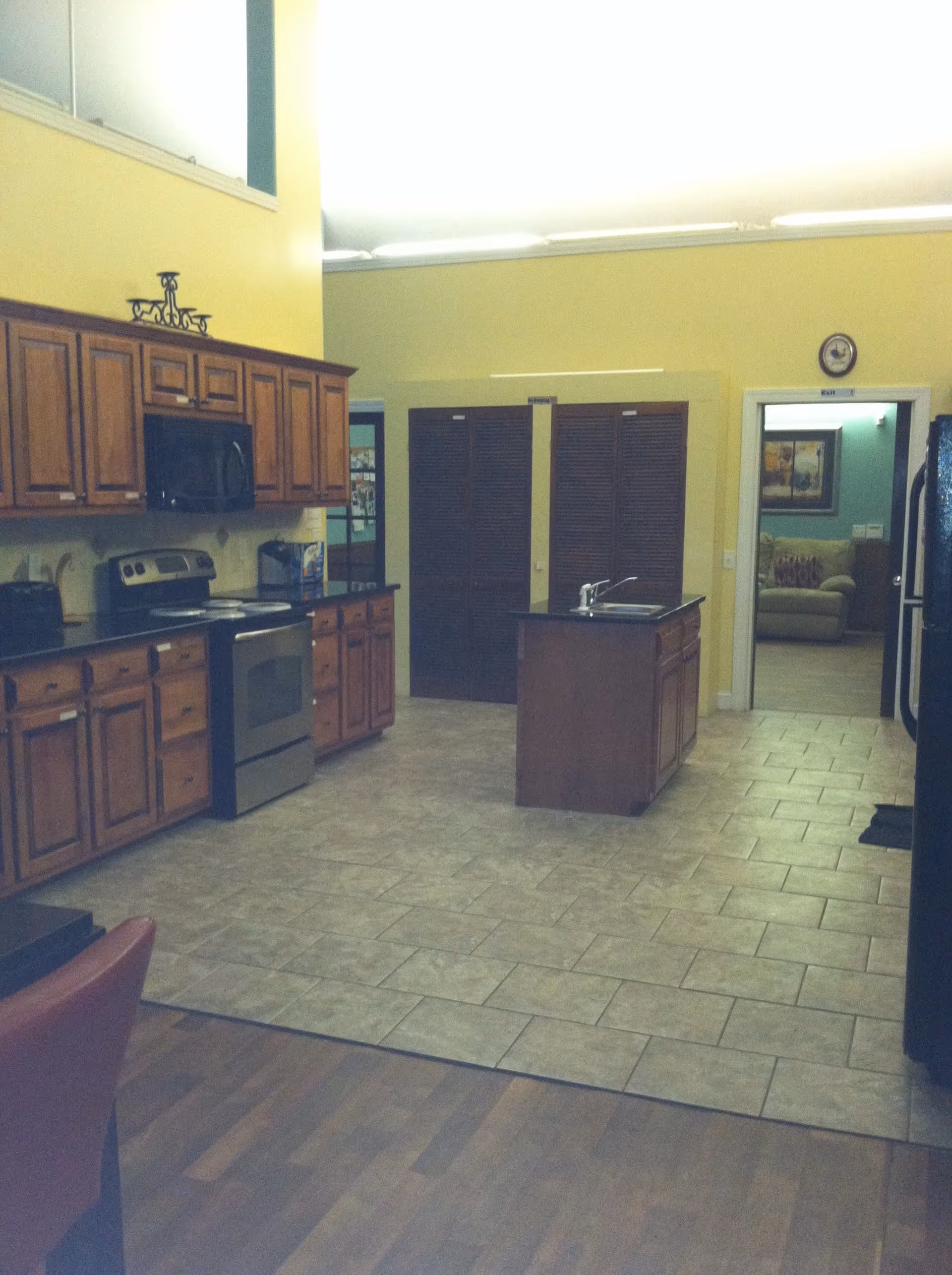 Interior view of a kitchen area with wooden cabinets, a stainless steel stove and microwave, a kitchen island with a sink, and tiled flooring. The walls are painted yellow, and there is an open doorway leading to a living room with a couch and framed artwork on the wall.