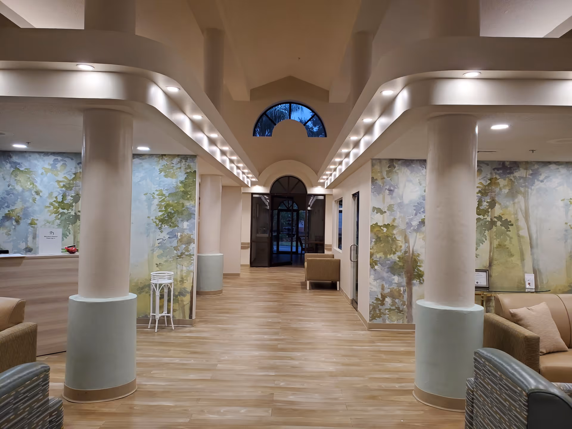 Interior view of a senior living facility hallway with wooden flooring, large white columns, and soft lighting along the ceiling edges. The walls feature nature-themed murals with trees and greenery. There are seating areas with beige and patterned chairs on both sides, and a glass door at the end of the hallway with an arched window above it.