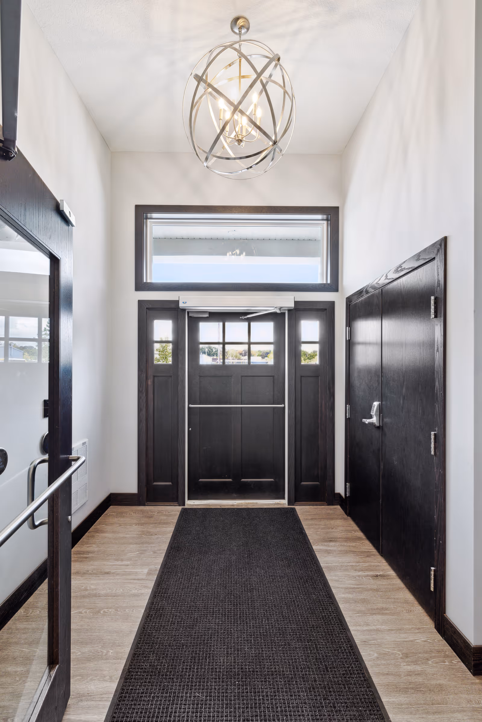 Entrance hallway with a black door featuring glass panels, a large window above the door, light wood flooring, a black floor mat, white walls, and a modern spherical chandelier hanging from the ceiling.