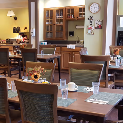Dining area with wooden tables and chairs, each table set with placemats, glasses, cups, and silverware. A small floral centerpiece is on one table. In the background, there is a kitchenette with wooden cabinets, a sink, and a clock on the wall.
