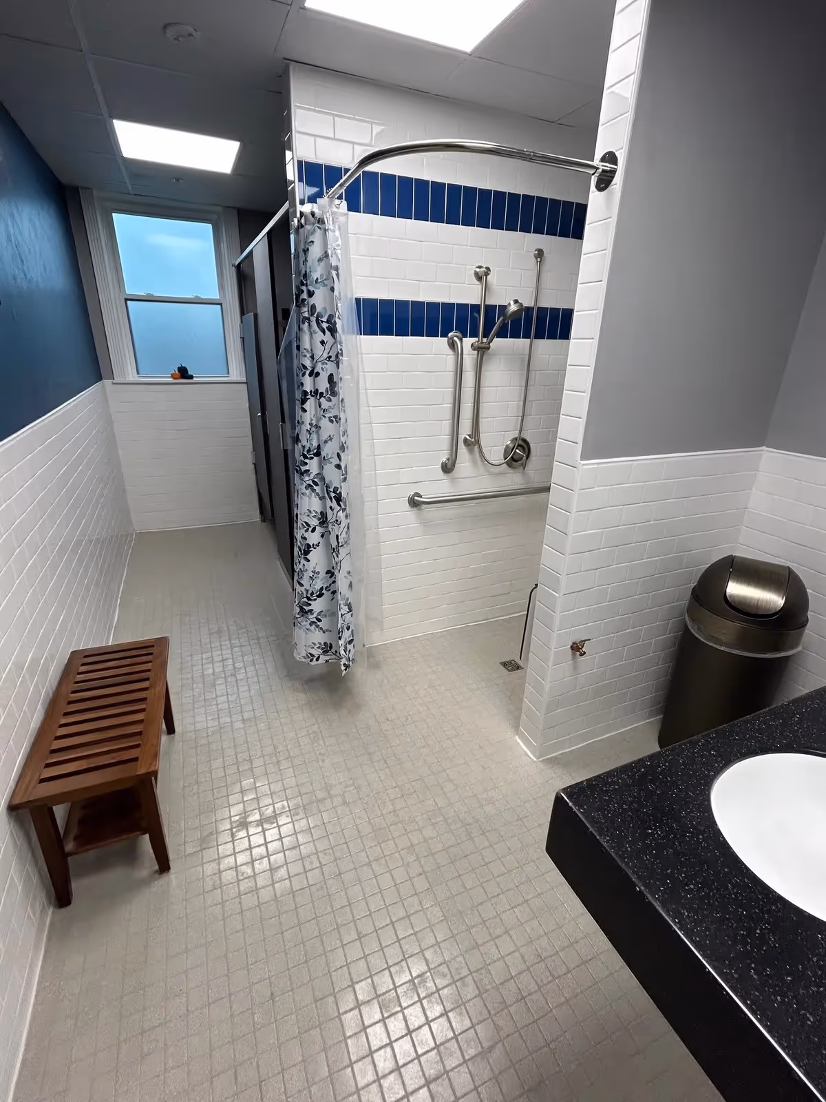 A clean, accessible bathroom with a walk-in shower featuring grab bars and a handheld showerhead. The shower has white tiles with two horizontal blue tile stripes. There is a wooden bench along one wall, a trash can near a black countertop with a white sink, and a window letting in natural light.