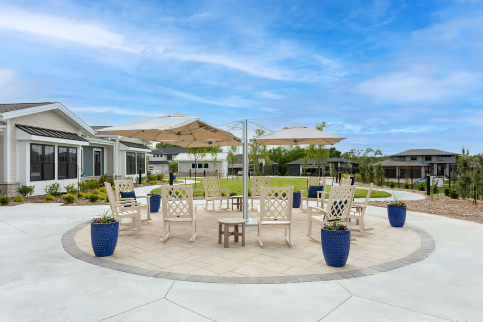 Outdoor seating area at CountryHouse Residence featuring a circular paved patio with beige chairs arranged around two large beige umbrellas. Blue planters with plants are placed around the seating area. The background shows a well-maintained lawn, trees, and residential-style buildings under a partly cloudy sky.