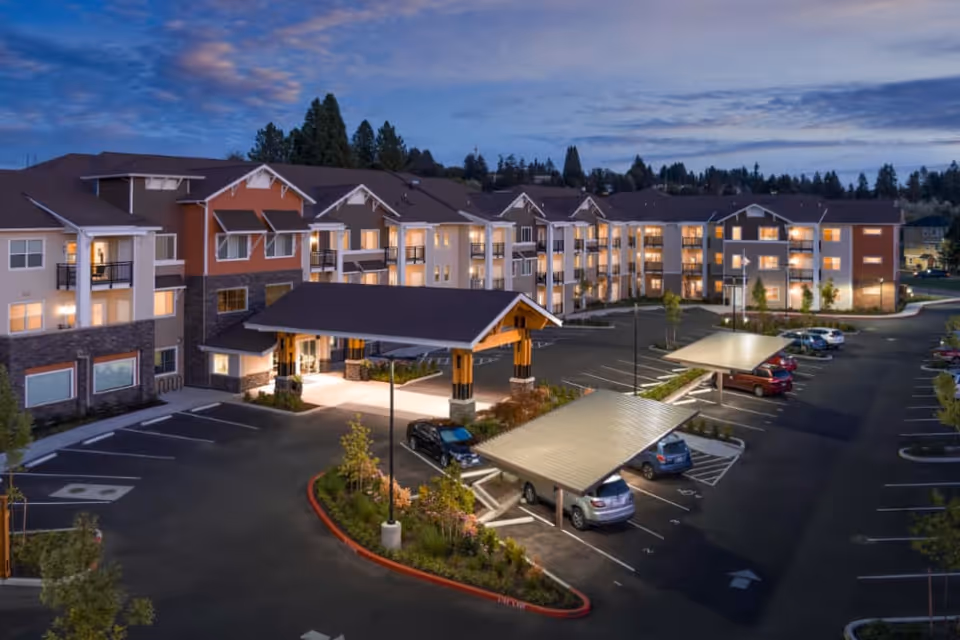 Exterior view of The Terraces At San Joaquin Gardens Village senior living facility at dusk, showing a large multi-story building with lit windows, covered parking spaces, landscaped areas, and a covered entrance.