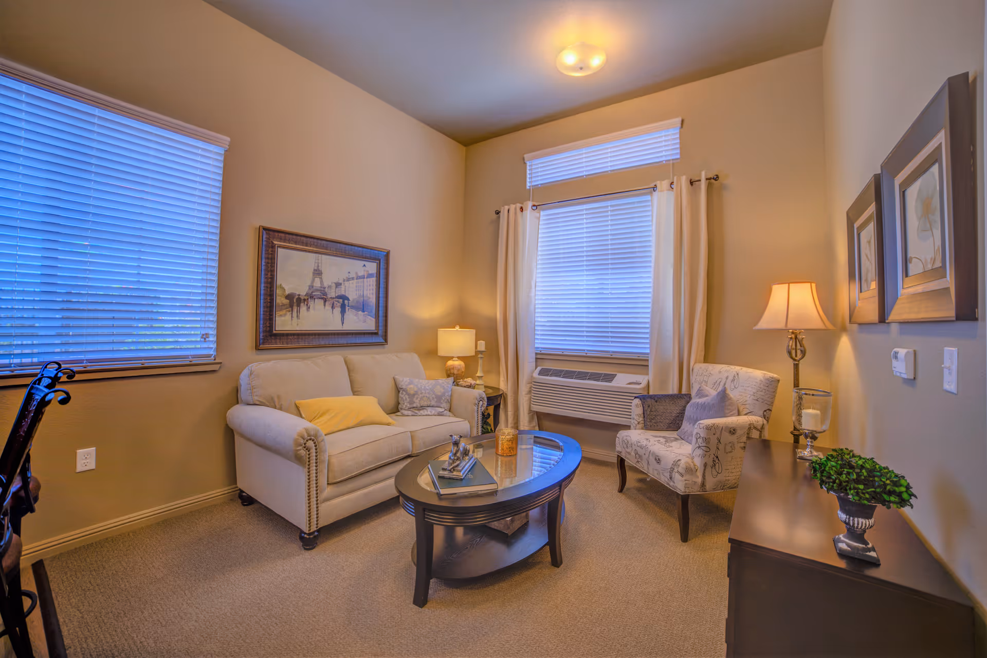 A cozy living room with a beige sofa adorned with pillows, a patterned armchair, a dark wooden coffee table with decorative items, two windows with blinds and curtains, a side table with a lamp, and framed artwork on the walls.
