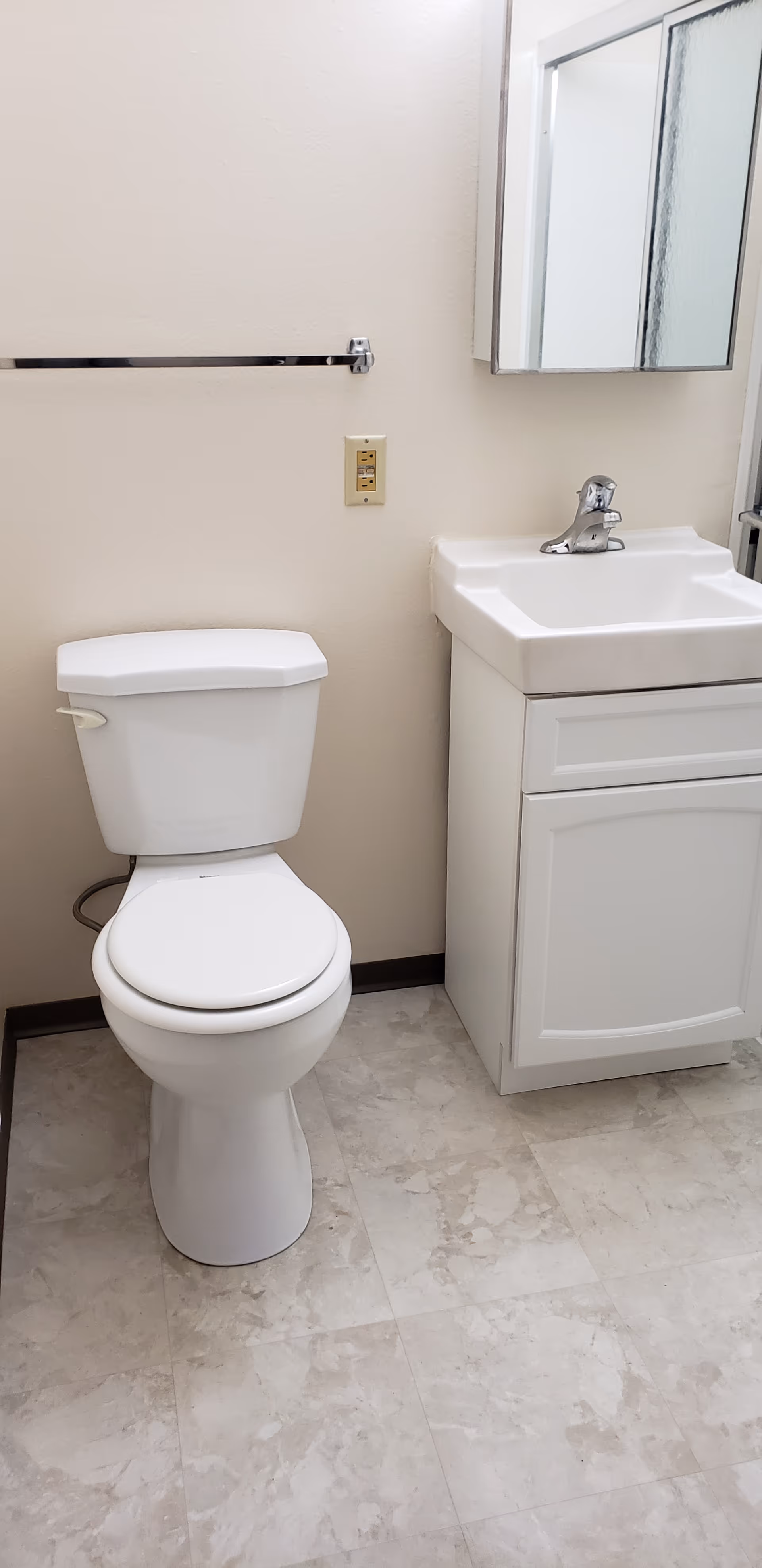 A small bathroom with a white toilet and a white sink cabinet. Above the sink is a mirrored medicine cabinet. There is a towel bar mounted on the wall above the toilet. The floor has light-colored tiles.