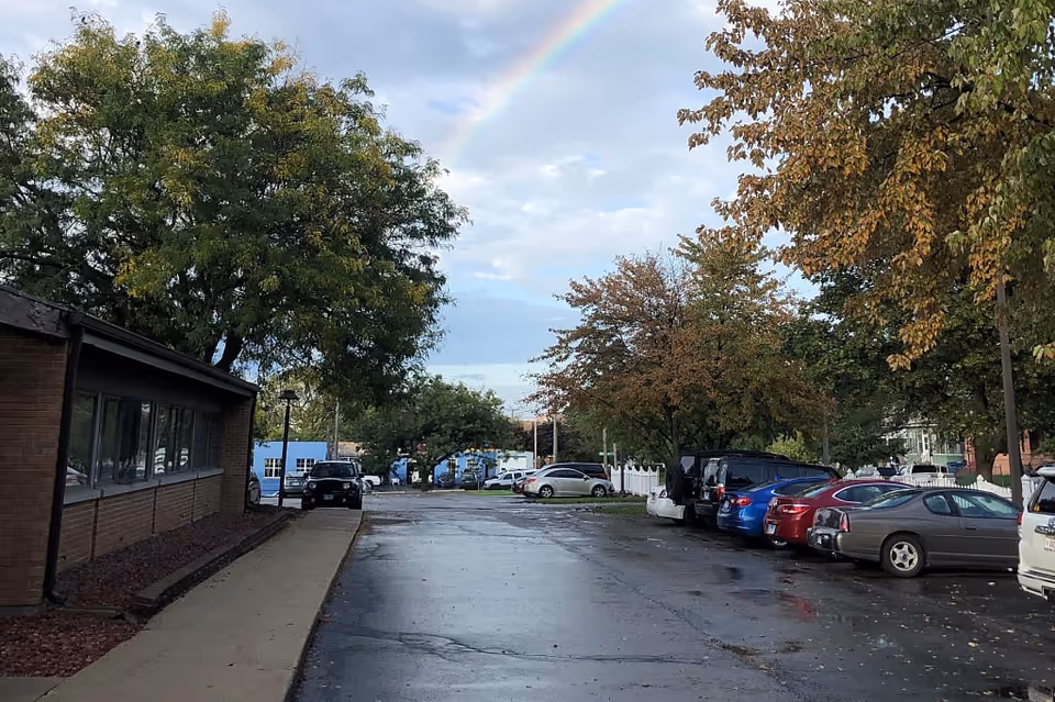 A parking lot next to a brick building with several parked cars and trees with green and autumn-colored leaves. A rainbow is visible in the sky above the scene.
