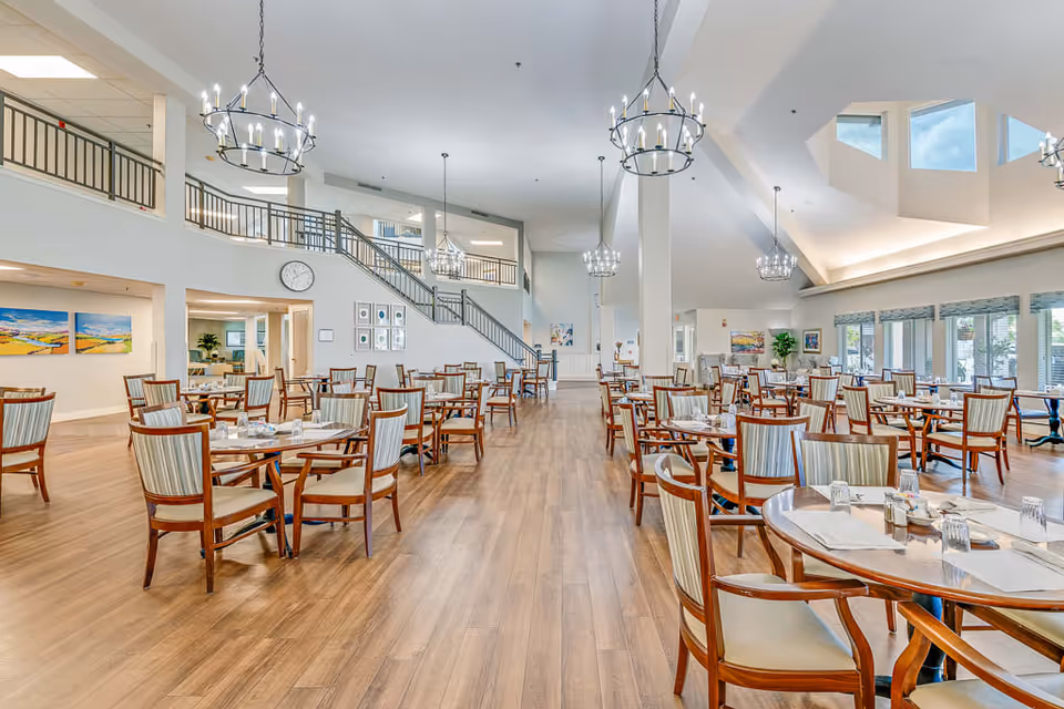 A spacious senior living dining room with multiple round wooden tables and chairs arranged neatly on a wooden floor. The room features high ceilings with large windows allowing natural light to fill the space. Modern chandeliers hang from the ceiling, and there is a staircase leading to an upper level. The walls are decorated with colorful paintings and a clock is visible near the staircase.