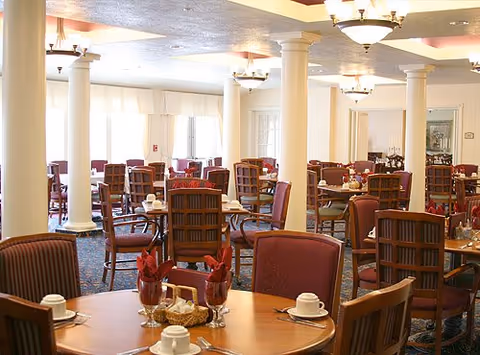 Spacious dining room with round wooden tables set for meals, upholstered chairs, and decorative columns.