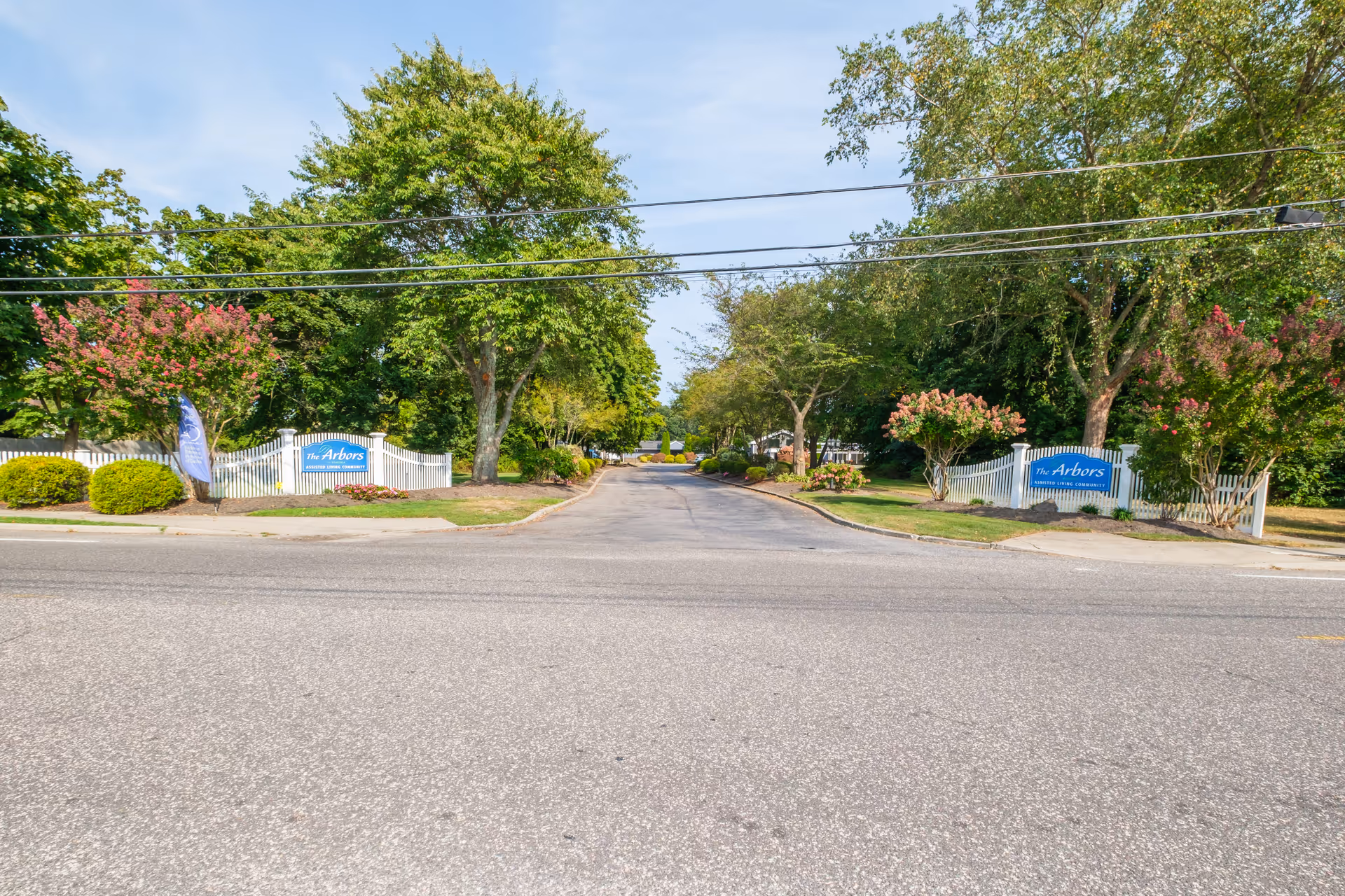 Entrance driveway to The Arbors senior living community flanked by white picket fences, trees, landscaping, and signage.