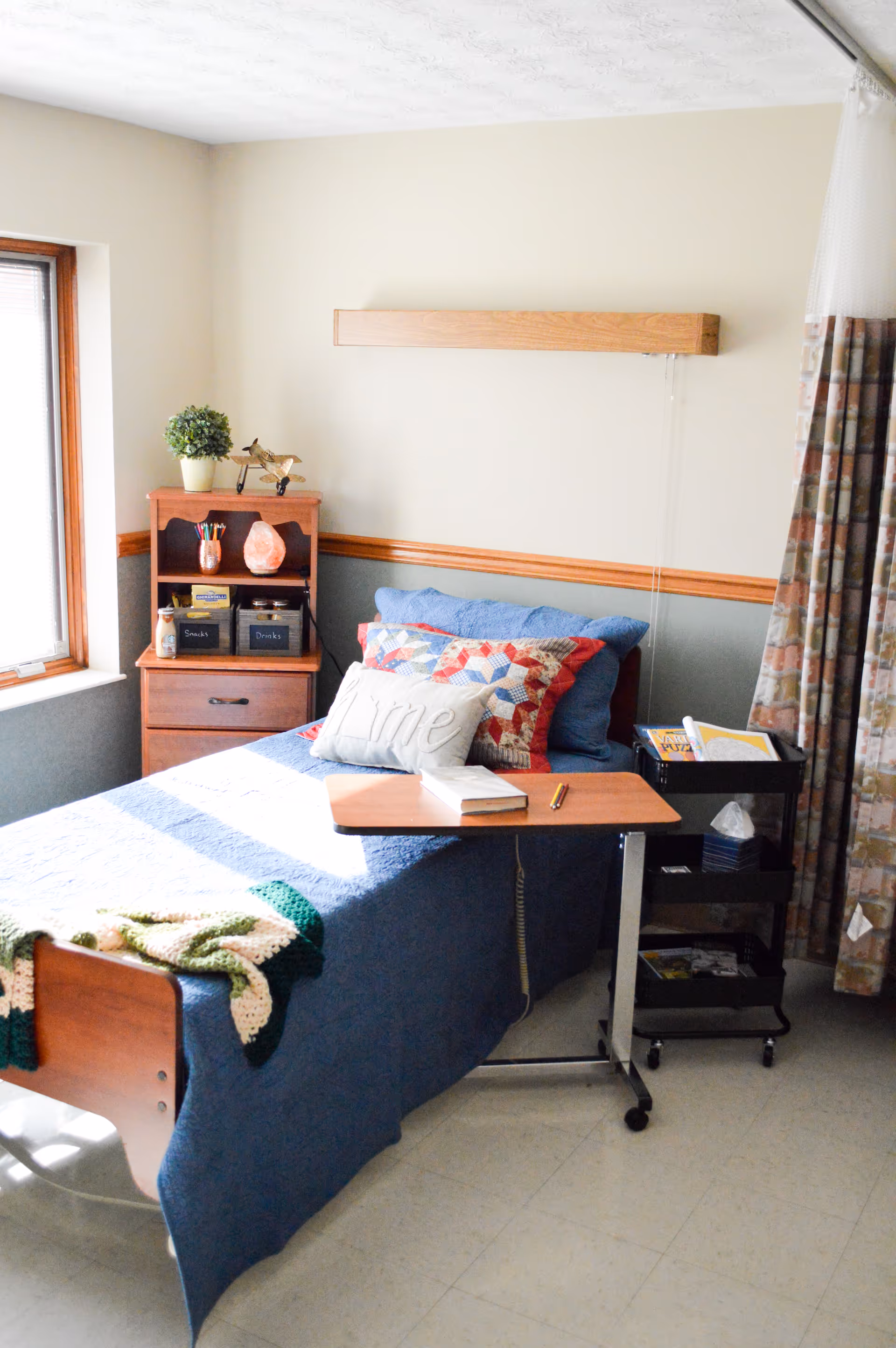 A single bed in a senior living facility room with a blue quilt, colorful pillows, and a small wooden overbed table with a book and pencil on it. Next to the bed is a small wooden nightstand with shelves holding snacks, drinks, and a potted plant. A rolling cart with tissues and magazines is beside the bed. The room has a window with natural light and patterned curtains.