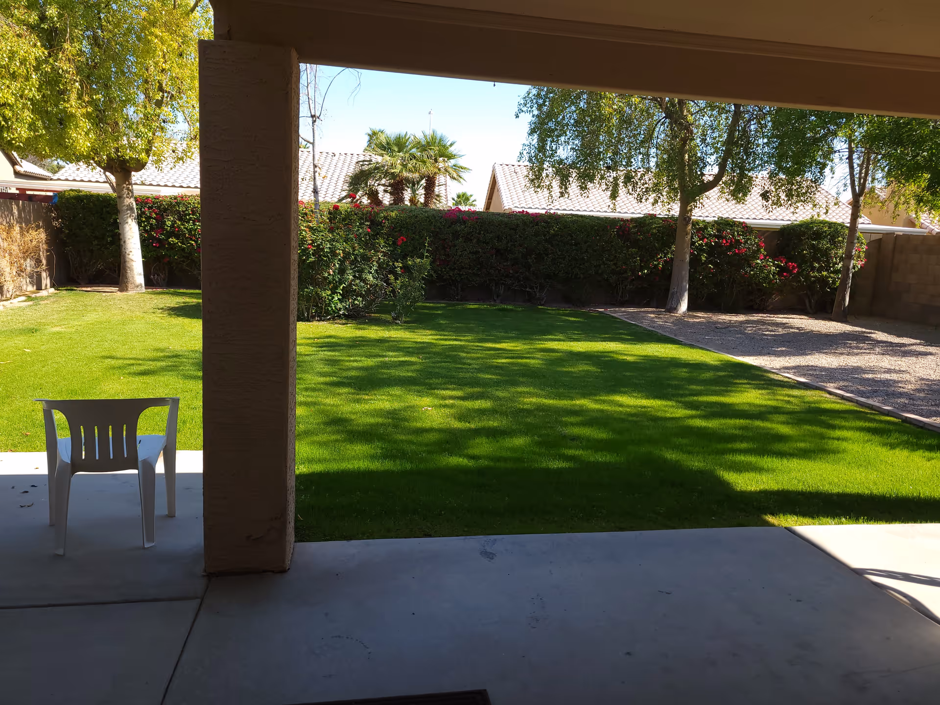 View from a covered patio looking out over a green lawn with a plastic chair, trees, and hedges under a clear sky.