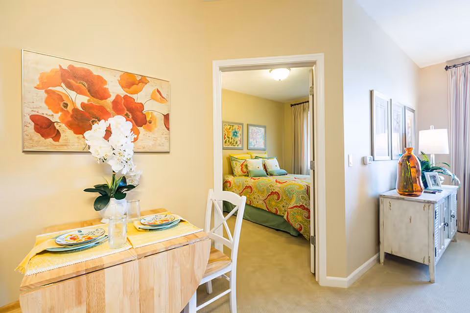 A cozy interior space featuring a small wooden dining table set for two with plates and a vase of white flowers. Adjacent to the dining area is a bedroom with a bed covered in a colorful paisley-patterned bedspread and decorative pillows. The walls are painted a soft beige, and the room is decorated with framed artwork and a large orange floral painting above the dining table. A sideboard with decorative items and a lamp is visible in the hallway leading to the bedroom.