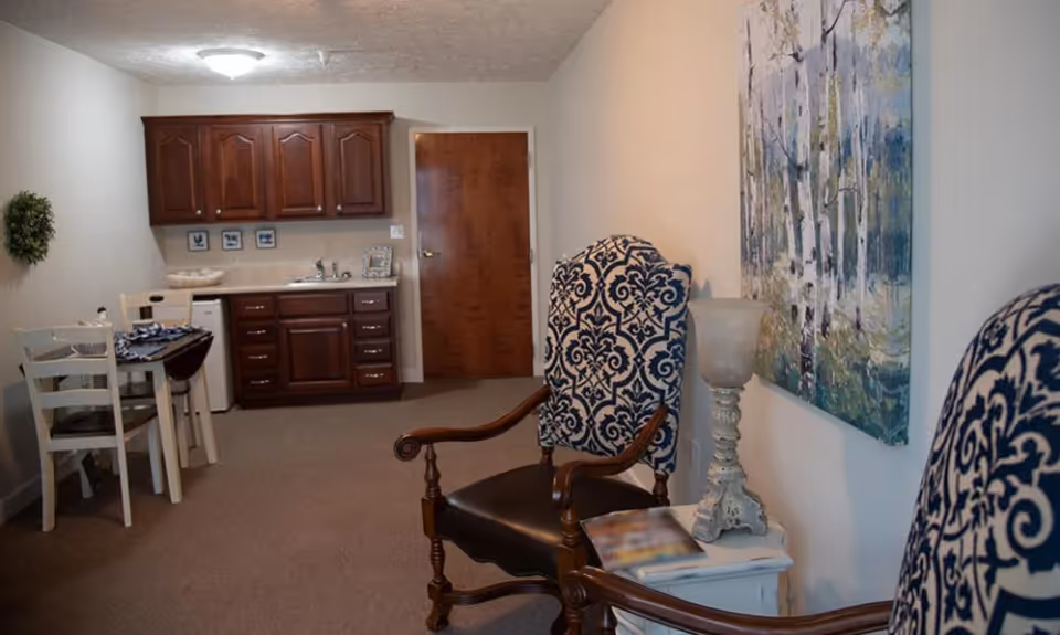 A cozy interior room with two patterned armchairs, a small white side table with a decorative lamp and a magazine, a wall painting of trees, a small dining table with two chairs, and a kitchenette with dark wooden cabinets, a sink, and a mini fridge. A closed wooden door is visible in the background.