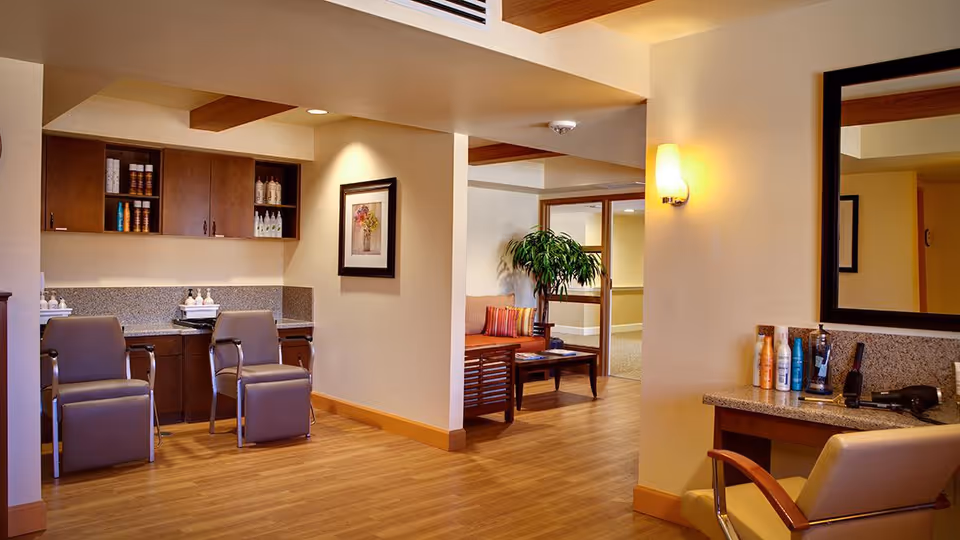 Interior view of a salon area in a senior living facility with two salon chairs in front of a counter with hair care products, a large mirror on the wall, and a seating area with a bench and a potted plant in the background.
