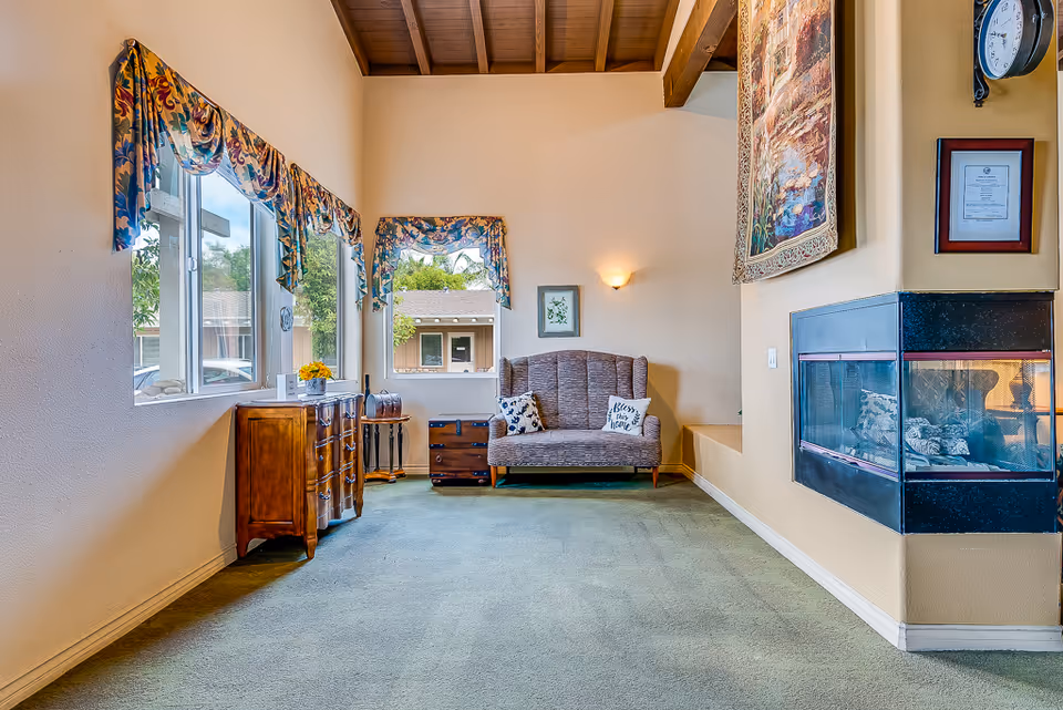 Sunlit common room with a sofa, decorative pillows, wooden cabinets, windows with floral valances, and a corner fireplace.