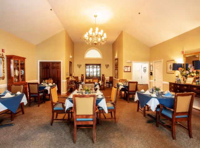 A warmly lit dining room with multiple tables covered in white and blue tablecloths, each set with plates, cups, and napkins. The room features wooden chairs with cushioned seats, a chandelier hanging from the ceiling, a wooden cabinet with glass doors, and framed artwork on the walls.