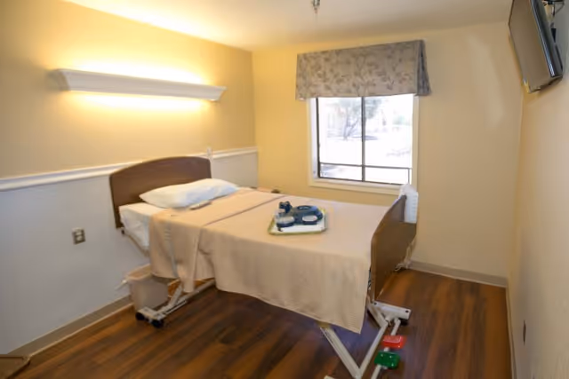 A simple patient bedroom with a single adjustable hospital bed, a tray with dishes on the bed, a window with a valance, wall lighting, and a wall-mounted TV.
