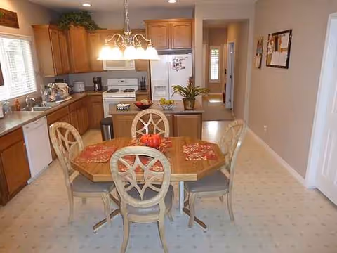 Dining area with a wooden table and chairs, kitchen cabinets and fridge in background.