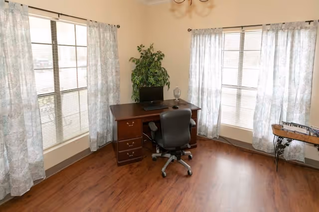Bright room with a wooden desk and office chair between two large curtained windows and a potted plant.