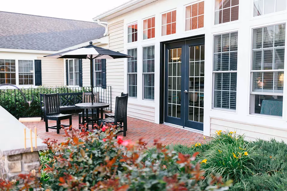 Outdoor patio area at Spring Arbor of Leesburg featuring a round table with an umbrella, surrounded by four chairs and a bench. The patio is paved with red bricks and bordered by greenery and flowering plants. The building exterior has beige siding, multiple windows with white frames, and a set of black double doors with glass panes.