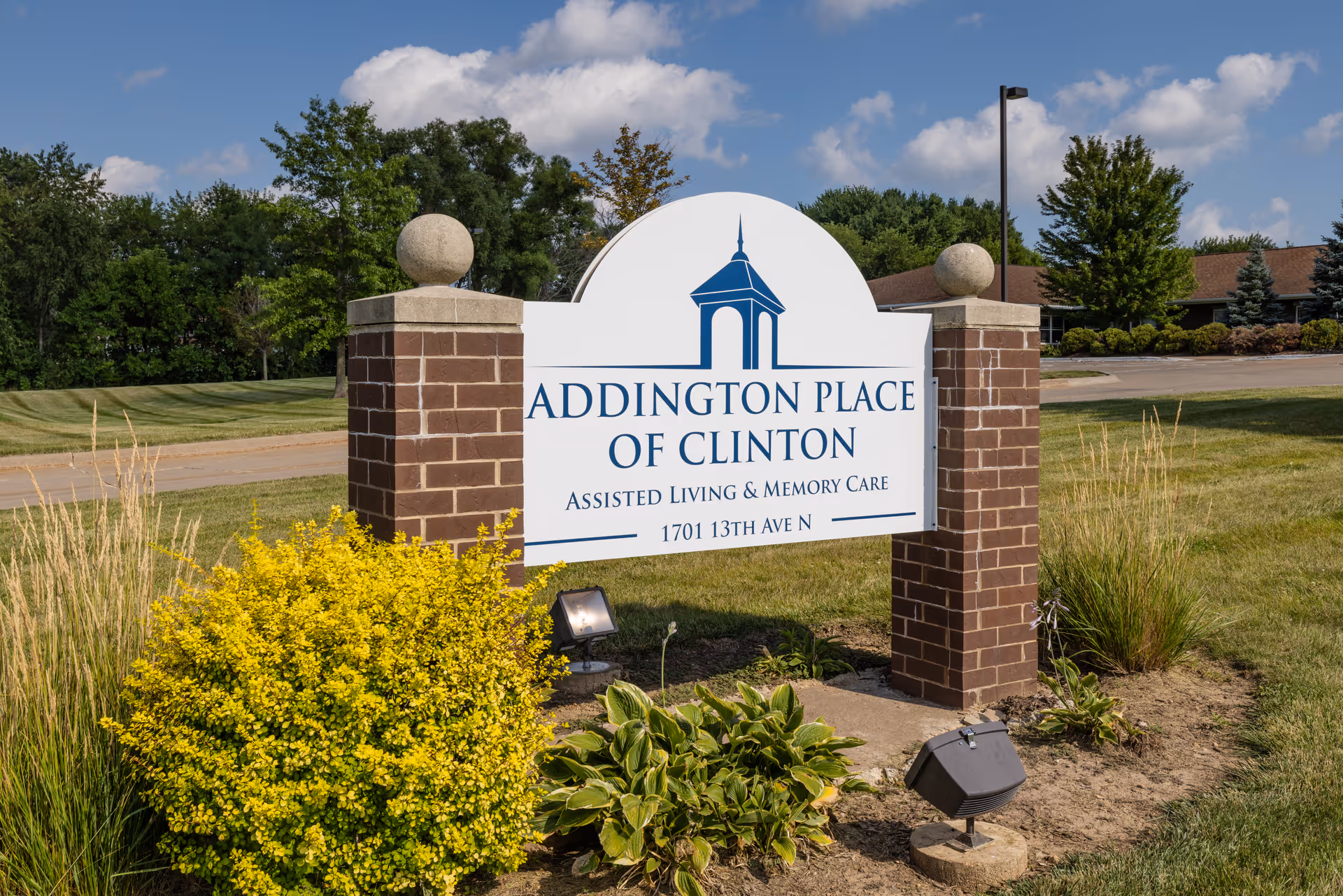 A brick-pillared roadside sign reading "Addington Place of Clinton Assisted Living & Memory Care" surrounded by landscaping with a building and trees in the background.