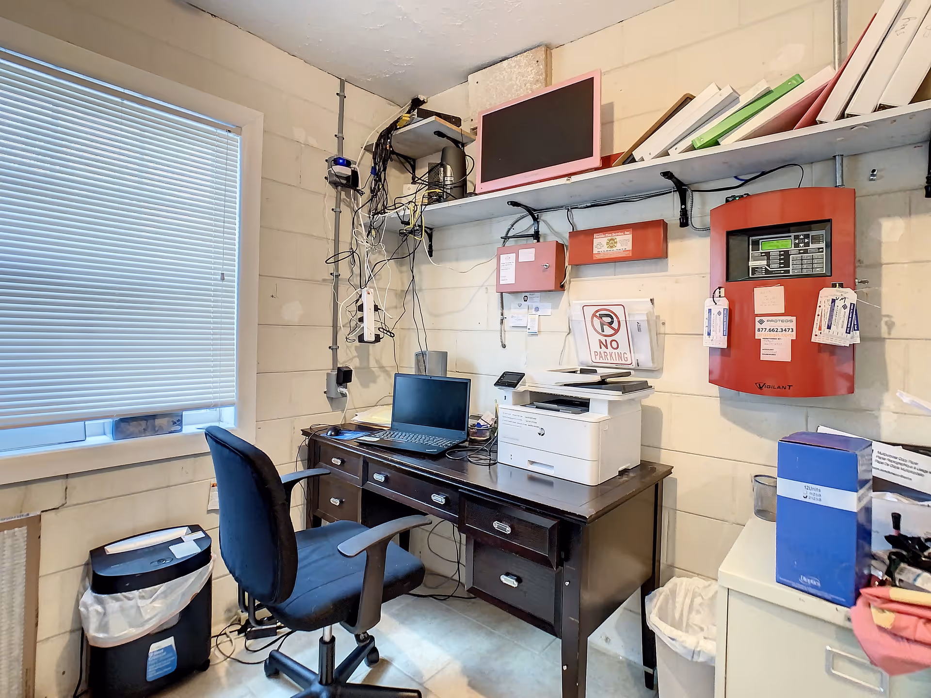 Small utility/office room with a desk holding a laptop and printer, an office chair, visible wiring and alarm panels on a cinderblock wall.