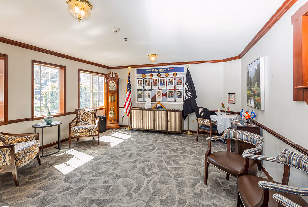 A well-lit sitting area with patterned armchairs and wooden chairs arranged around the room. There is a grandfather clock near the windows, a small table with a decorative vase, and a cabinet against the far wall displaying a 'Wall of Honor' with photos and flags, including the American flag and a POW/MIA flag. The room has carpeted flooring, white walls with wooden trim, and framed artwork on the walls.