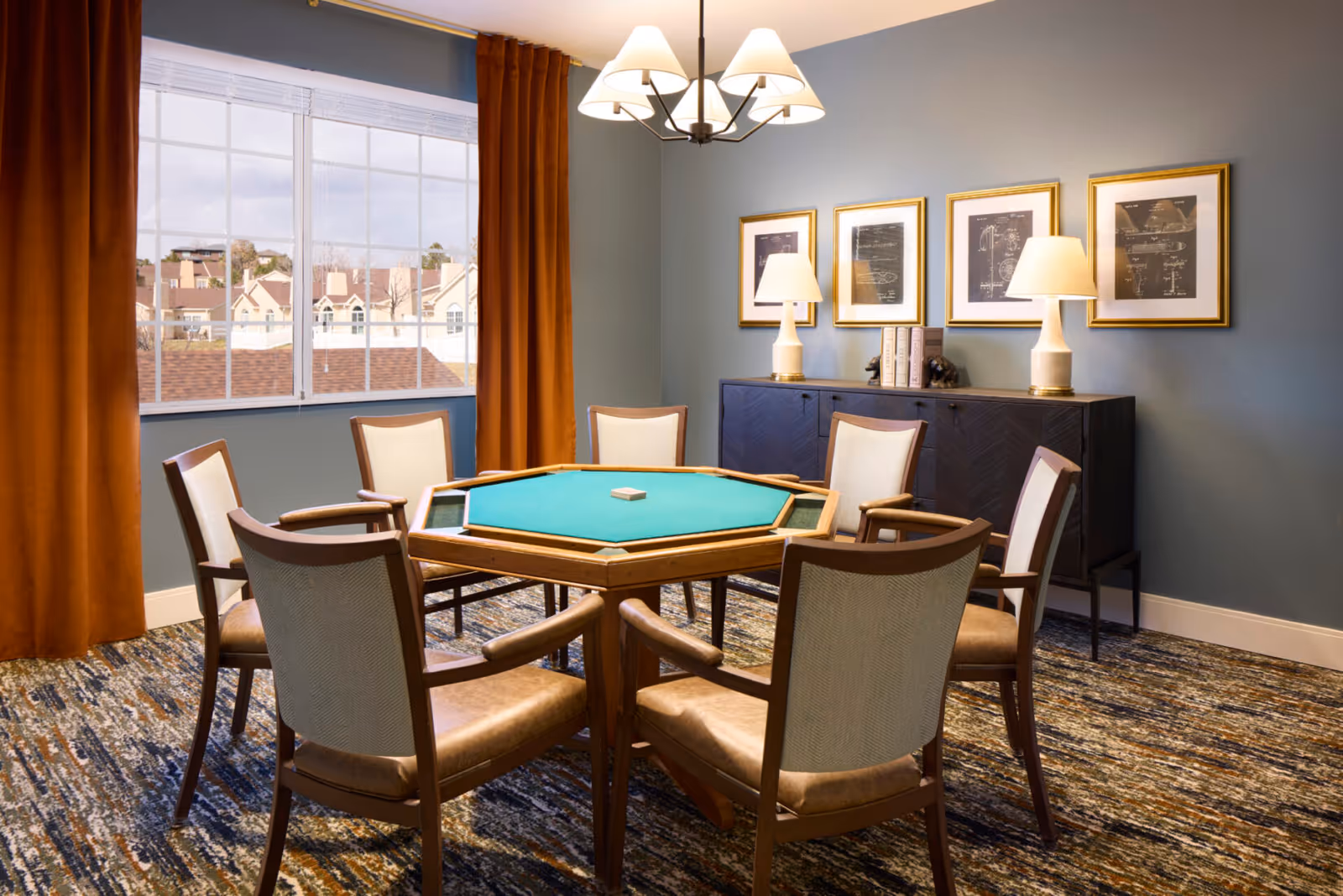 A cozy interior game room with a hexagonal card table surrounded by six chairs, a sideboard with lamps and framed art, and a large window with orange curtains.