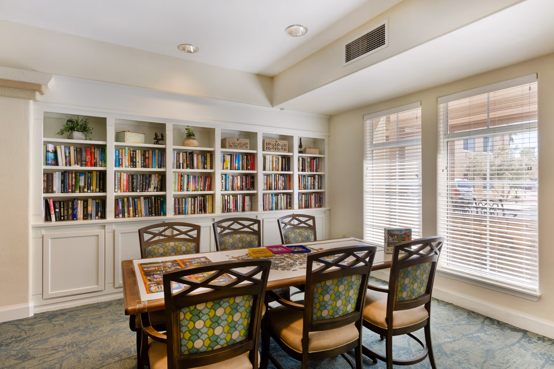 Well-lit communal library/activity room with built-in bookshelves, a table surrounded by six chairs, and large windows with blinds.