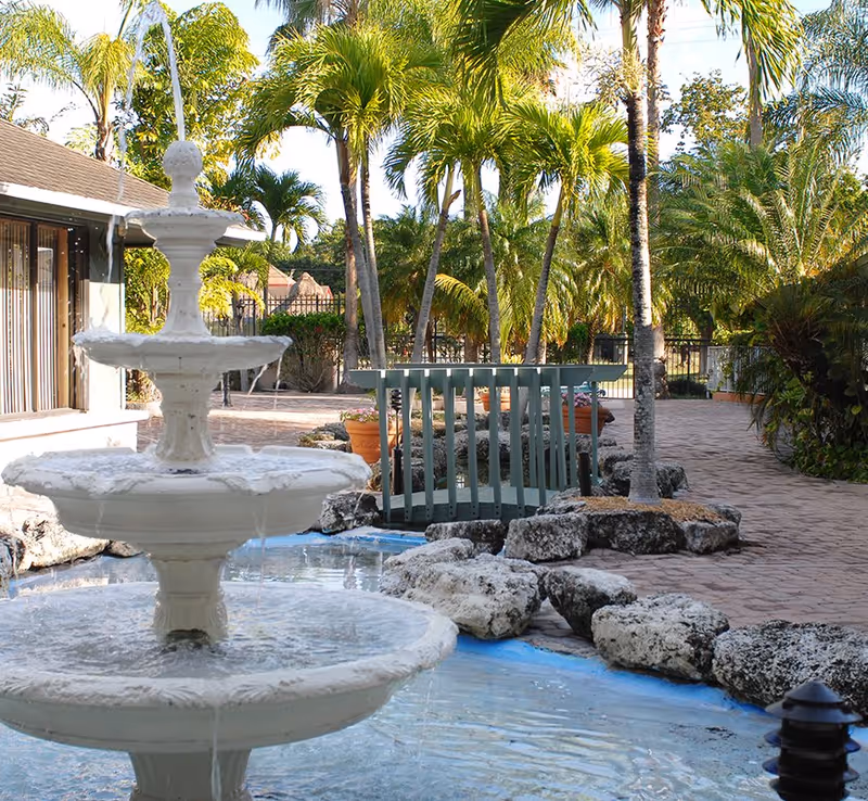 A three-tier white fountain in a landscaped courtyard with palm trees, rocks, and a small footbridge.