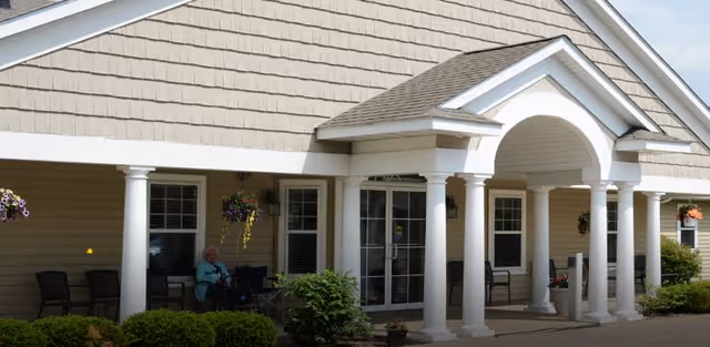Exterior view of a senior living facility entrance with white columns, a covered porch, and beige siding. There are chairs along the wall and a person sitting near the entrance. Hanging flower baskets and shrubs decorate the area.