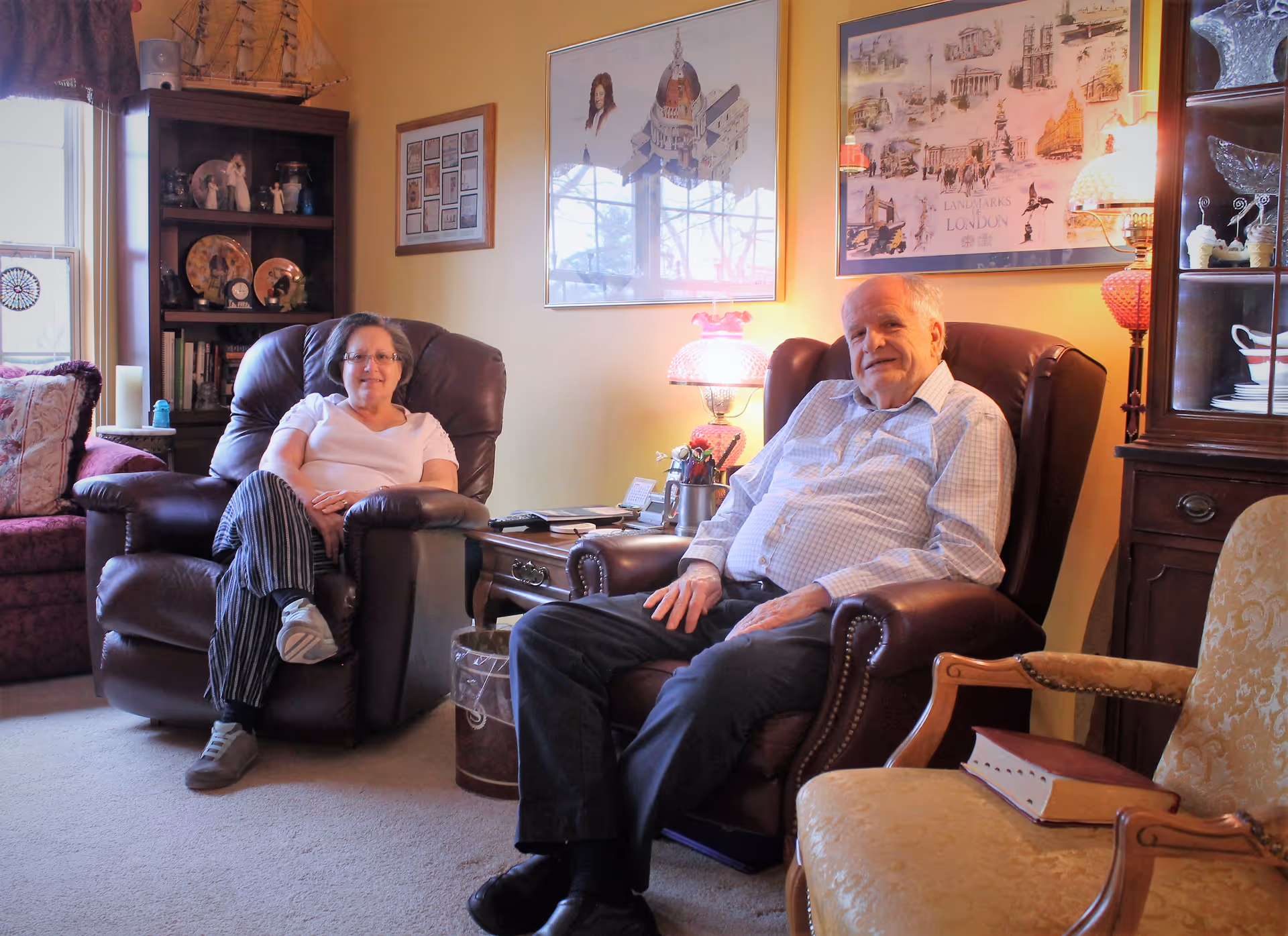 An elderly man and woman sitting comfortably in leather armchairs in a warmly lit living room. The room features framed artwork on the walls, a wooden bookshelf with decorative items, a side table with a lamp, and a cushioned armchair with a book on it.