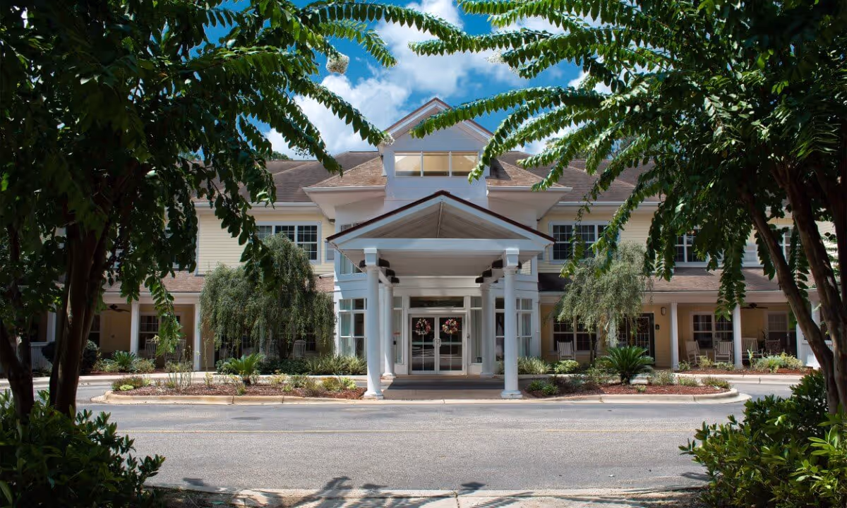 The front entrance of a senior living building with a covered porte-cochere, glass doors, and landscaped driveway framed by trees.