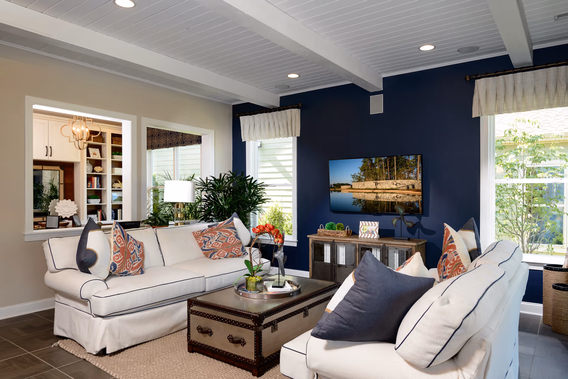 Bright furnished living room with white sofas, patterned pillows, a navy accent wall with mounted TV, coffee table, and large windows.