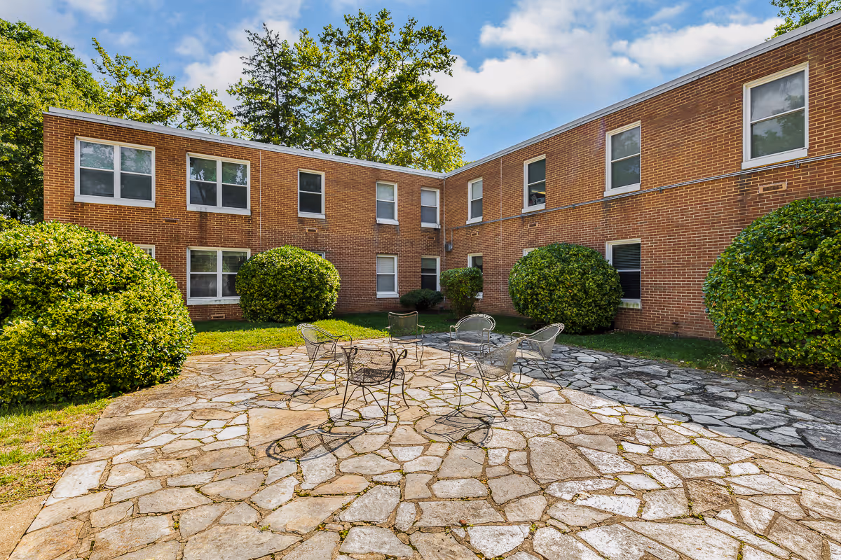 Outdoor courtyard area with stone paving, surrounded by a two-story brick building with multiple windows. There are several metal chairs arranged in a circle on the stone patio, and large green bushes and trees around the perimeter under a partly cloudy sky.