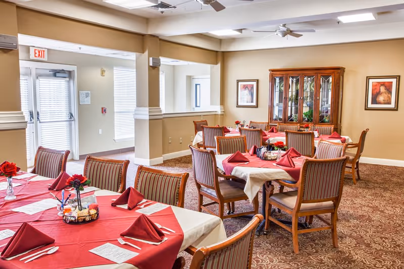 Bright dining room with multiple tables set with red tablecloths and folded napkins, wooden chairs, and a china cabinet against the far wall.
